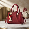 A structured red handbag with a woven chevron pattern, dual top handles, a detachable shoulder strap, and a decorative pom-pom tassel charm, displayed on a marble table with a gold cylinder and vase in the background.