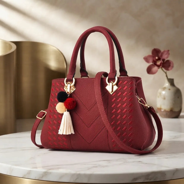 A structured red handbag with a woven chevron pattern, dual top handles, a detachable shoulder strap, and a decorative pom-pom tassel charm, displayed on a marble table with a gold cylinder and vase in the background.