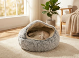 A fluffy gray round pet bed with a cream interior, shown on a rug in a sunlit room with a cat sleeping inside. A potted plant and chair are visible in the background.