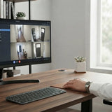A modern home office setup with a large multi-view security monitor showing multiple camera feeds, alongside a wireless keyboard and mouse on a wooden desk. A person's hand and arm are visible on the desk, and the scene is set against a bright, neutral background with a window.