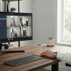 A modern home office setup with a large multi-view security monitor showing multiple camera feeds, alongside a wireless keyboard and mouse on a wooden desk. A person's hand and arm are visible on the desk, and the scene is set against a bright, neutral background with a window.