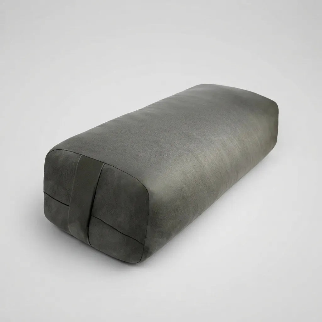 A dark gray rectangular yoga prop pillow with a textured fabric surface and a strap detail on one end, shown on a light gray background.