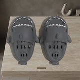 A pair of dark gray shark-shaped clogs with white teeth details, displayed on a light beige stone surface.