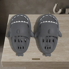 A pair of dark gray shark-shaped clogs with white teeth details, displayed on a light beige stone surface.