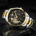 A two-tone stainless steel and gold wristwatch with a black dial, featuring a skeletonized center, dual subdials, and a gold-tone metal bracelet, shown on a dark reflective surface.
