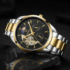 A two-tone stainless steel and gold wristwatch with a black dial, featuring a skeletonized center, dual subdials, and a gold-tone metal bracelet, shown on a dark reflective surface.
