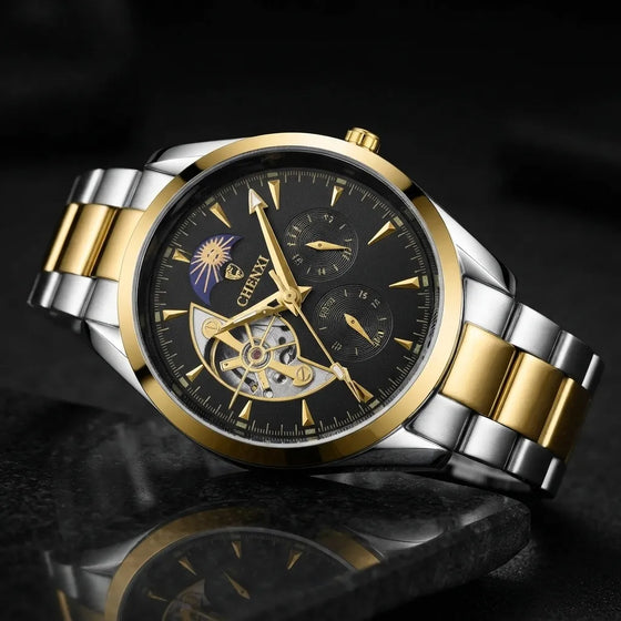 A two-tone stainless steel and gold wristwatch with a black dial, featuring a skeletonized center, dual subdials, and a gold-tone metal bracelet, shown on a dark reflective surface.