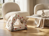 A small fabric pet play tent with a cherry and polka dot pattern sits on a light wood floor in a modern living room setting with beige armchairs and a marble coffee table.