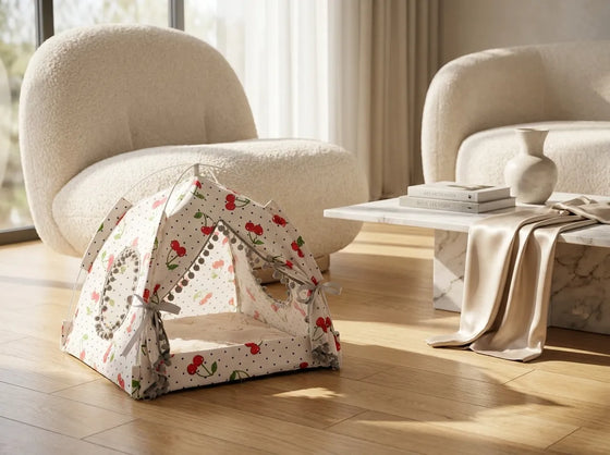A small fabric pet play tent with a cherry and polka dot pattern sits on a light wood floor in a modern living room setting with beige armchairs and a marble coffee table.