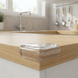 A clear plastic corner guard bumper attached to the edge of a light oak kitchen countertop in a bright, modern kitchen setting.