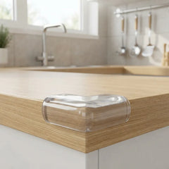 A clear plastic corner guard bumper attached to the edge of a light oak kitchen countertop in a bright, modern kitchen setting.