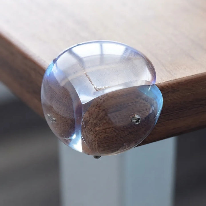A close-up of a clear acrylic table corner protector with a dark brown wood backing, mounted on a wooden table corner.