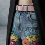 Close-up of a person wearing blue floral-print jeans with a pink belt, a black coat draped over the shoulders, and a gray concrete wall in the background.