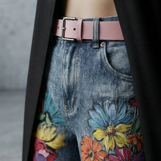 Close-up of a person wearing blue floral-print jeans with a pink belt, a black coat draped over the shoulders, and a gray concrete wall in the background.