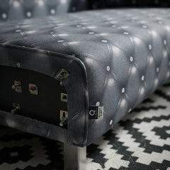 Close-up of a tufted upholstered bench or ottoman in dark gray fabric with a diamond button pattern, shown on a black-and-white geometric patterned rug.