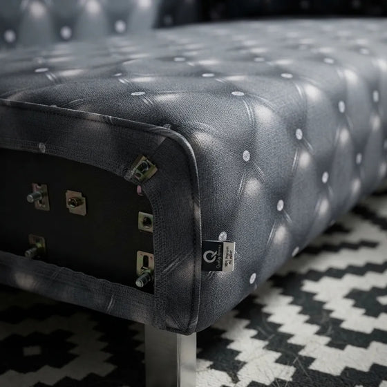Close-up of a tufted upholstered bench or ottoman in dark gray fabric with a diamond button pattern, shown on a black-and-white geometric patterned rug.