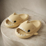 A pair of pale yellow shark-shaped slide slippers with white teeth and black eyes, displayed on a round marble pedestal against a textured beige wall.