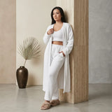 A woman stands indoors against a textured stone column wearing a white lounge set consisting of a cropped knit top, matching wide-leg pants, a long open-front cardigan, and beige slippers. A dark vase with a dried palm leaf sits to the left.