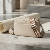 A cream-colored pebbled leather crossbody bag with a patterned brown and beige strap, resting on a marble surface next to a rolled cream fabric.