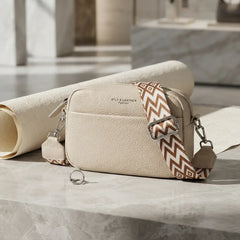 A cream-colored pebbled leather crossbody bag with a patterned brown and beige strap, resting on a marble surface next to a rolled cream fabric.