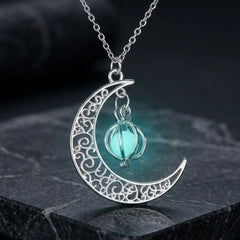 A silver crescent moon pendant necklace with intricate filigree detailing and a hanging light blue glass orb, displayed on a dark textured stone surface.