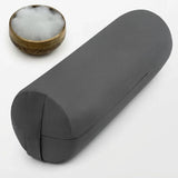 A dark gray cylindrical yoga bolster pillow shown on a white background, with a small brass bowl containing white cotton padding in the upper left corner.
