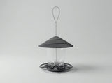 A modern bird feeder with a dark gray ribbed conical roof, a clear cylindrical body, and a circular base with a hanging loop, shown on a light gray background.