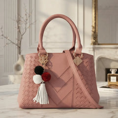 A dusty pink woven leather handbag with double top handles, a shoulder strap, and decorative pom-pom tassels, displayed on a marble surface in an elegant interior setting.