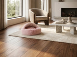 A pink fuzzy pet bed placed on a cream rug in a modern living room with hardwood floors, a beige armchair, and a marble coffee table.