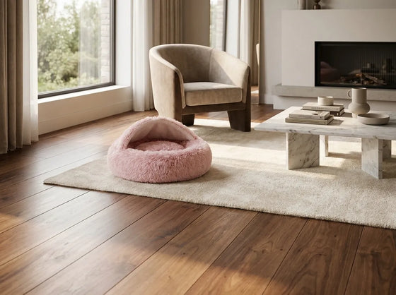 A pink fuzzy pet bed placed on a cream rug in a modern living room with hardwood floors, a beige armchair, and a marble coffee table.