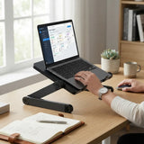 A black adjustable laptop stand holding an open laptop on a wooden desk, with a person's hands using the trackpad in a bright home office setting.