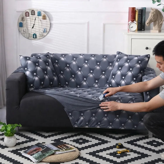 A gray tufted sofa slipcover with matching back cushions is being fitted onto a dark gray sofa in a living room. A person is kneeling on a black-and-white patterned rug, using a small tool to smooth the cover.