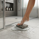 A close-up of a person's foot with soap lather pressing down on a gray and white scrubbing brush on a tiled bathroom floor in front of a glass shower door.