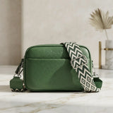A green pebbled leather crossbody bag with a wide, patterned black and white chevron strap, resting on a light marble surface.