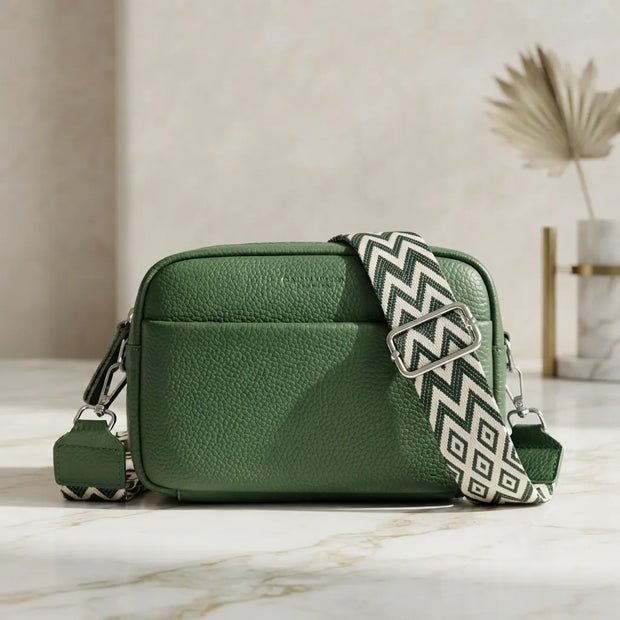 A green pebbled leather crossbody bag with a wide, patterned black and white chevron strap, resting on a light marble surface.