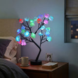 A black LED tree light with glowing multicolor rose-shaped bulbs placed on a wooden bedside table next to a bed in a dimly lit bedroom.