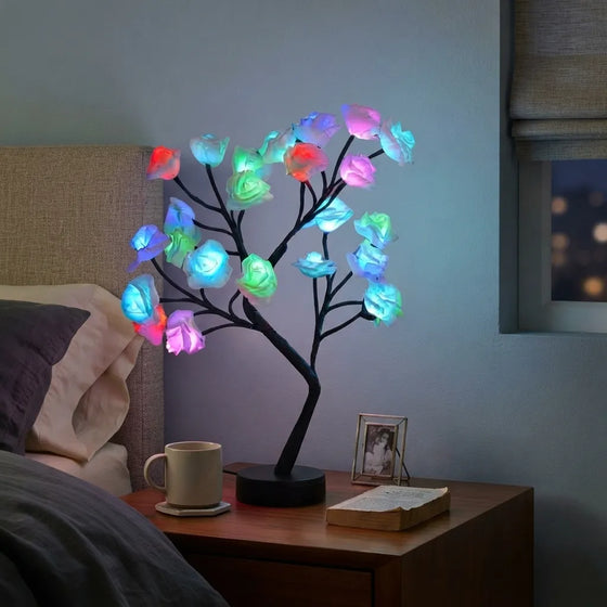 A black LED tree light with glowing multicolor rose-shaped bulbs placed on a wooden bedside table next to a bed in a dimly lit bedroom.