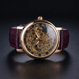 A luxury skeleton watch with a rose gold-toned case, intricate gold-tone mechanical gears visible through the transparent dial, and a deep burgundy leather strap, set against a dark reflective background.