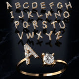 Gold letter A initial ring set with pavé crystals, shown alongside a matching gold ring with a round-cut crystal. Above them, matching gold alphabet letters from A to Z are arranged on a dark reflective background.