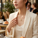 A woman in a cream-colored blazer and top wears a delicate gold necklace with a sunburst flower pendant, sitting at a cafe table with croissants and coffee cups in the background.