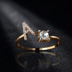 A gold ring featuring a letter 'A' adorned with small clear stones on one side and a large round clear center stone on the other, displayed on a dark reflective surface.