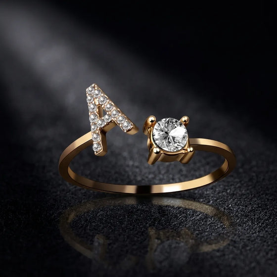 A gold ring featuring a letter 'A' adorned with small clear stones on one side and a large round clear center stone on the other, displayed on a dark reflective surface.