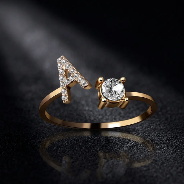 A gold ring featuring a letter 'A' adorned with small clear stones on one side and a large round clear center stone on the other, displayed on a dark reflective surface.