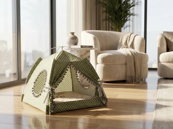 A green and white polka dot fabric pet tent with pom-pom trim, set on a wooden floor in a bright, modern living room with beige armchairs and large windows.