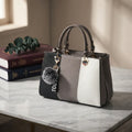 A structured handbag with a color-blocked design in black, gray, and white, featuring dual top handles, a gray pom-pom charm, and a heart-shaped metal accent, displayed on a marble tabletop next to stacked books.
