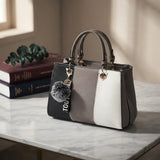 A structured handbag with a color-blocked design in black, gray, and white, featuring dual top handles, a gray pom-pom charm, and a heart-shaped metal accent, displayed on a marble tabletop next to stacked books.