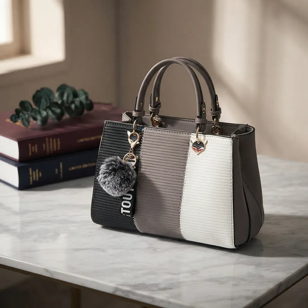 A structured handbag with a color-blocked design in black, gray, and white, featuring dual top handles, a gray pom-pom charm, and a heart-shaped metal accent, displayed on a marble tabletop next to stacked books.