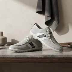 A pair of light gray knit lace-up sneakers with white and black accents, displayed on a marble countertop in a modern, minimalist room setting.