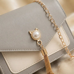 A close-up of a light gray and cream faux leather handbag with a gold chain strap and a decorative gold tassel clasp featuring a white faux pearl cat head.