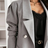 A close-up of a person wearing a light gray double-breasted blazer with dramatic puffed sleeves, layered over a black lace-trim camisole and a black belt with a gold-tone V-shaped buckle.